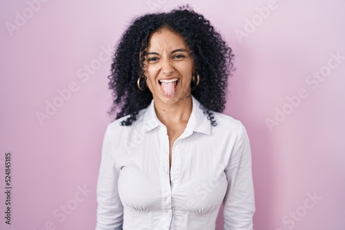 Hispanic woman with curly hair standing over pink background sticking tongue out happy with funny expression. emotion concept.