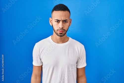 Young hispanic man standing over blue background skeptic and nervous, frowning upset because of problem. negative person.