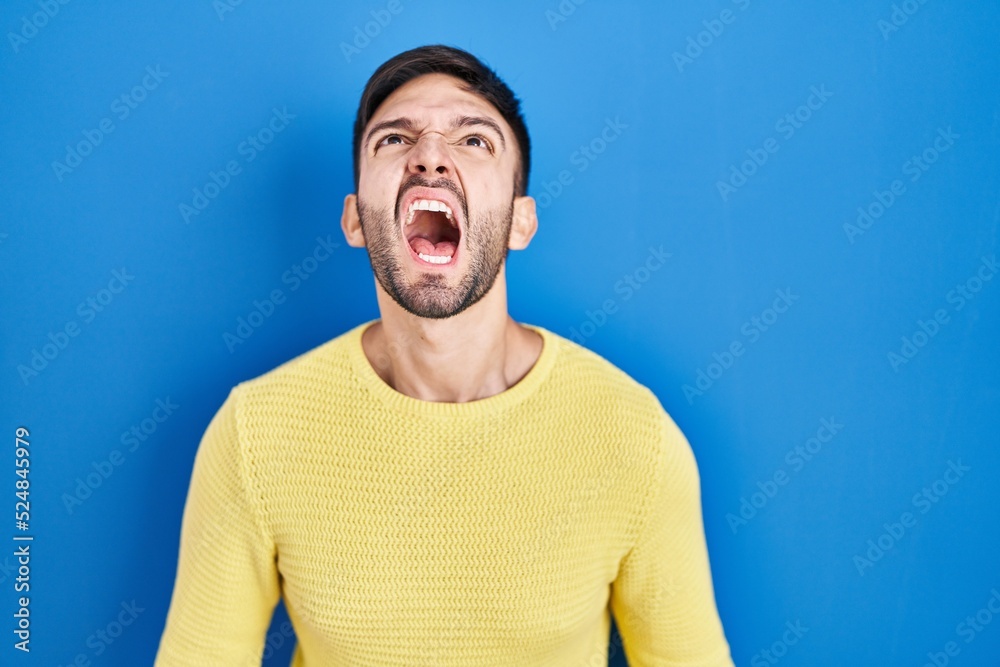Hispanic man standing over blue background angry and mad screaming frustrated and furious, shouting with anger. rage and aggressive concept. Hispanic man standing over blue background angry and mad screaming frustrated and furious, shouting with anger. rage and aggressive concept.