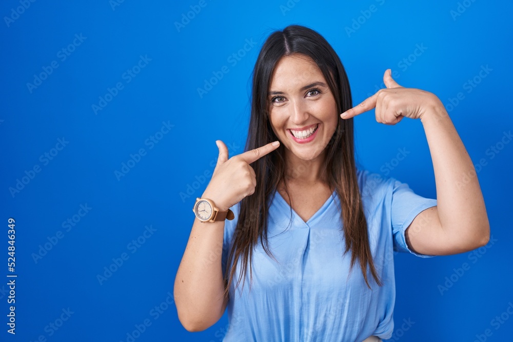 Young brunette woman standing over blue background smiling cheerful showing and pointing with fingers teeth and mouth. dental health concept.