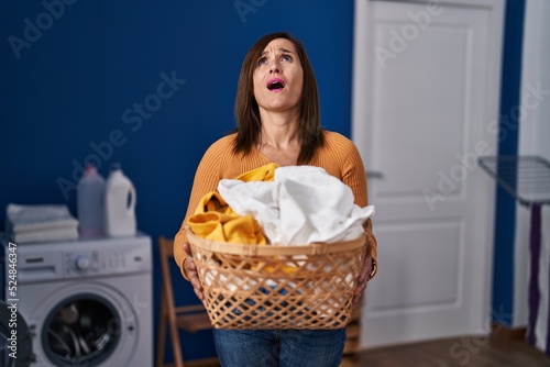 Middle age woman holding laundry basket at laundry room angry and mad screaming frustrated and furious, shouting with anger looking up.