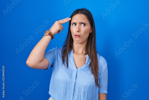 Young brunette woman standing over blue background shooting and killing oneself pointing hand and fingers to head like gun  suicide gesture.