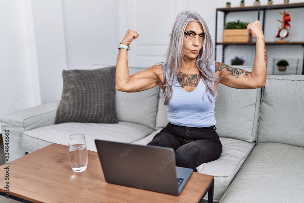 Middle age grey-haired woman using laptop at home showing arms muscles smiling proud. fitness concept.