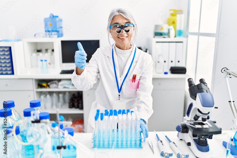 Middle age grey-haired woman wearing scientist uniform smiling happy and positive, thumb up doing excellent and approval sign Middle age grey-haired woman wearing scientist uniform smiling happy and positive, thumb up doing excellent and approval sign