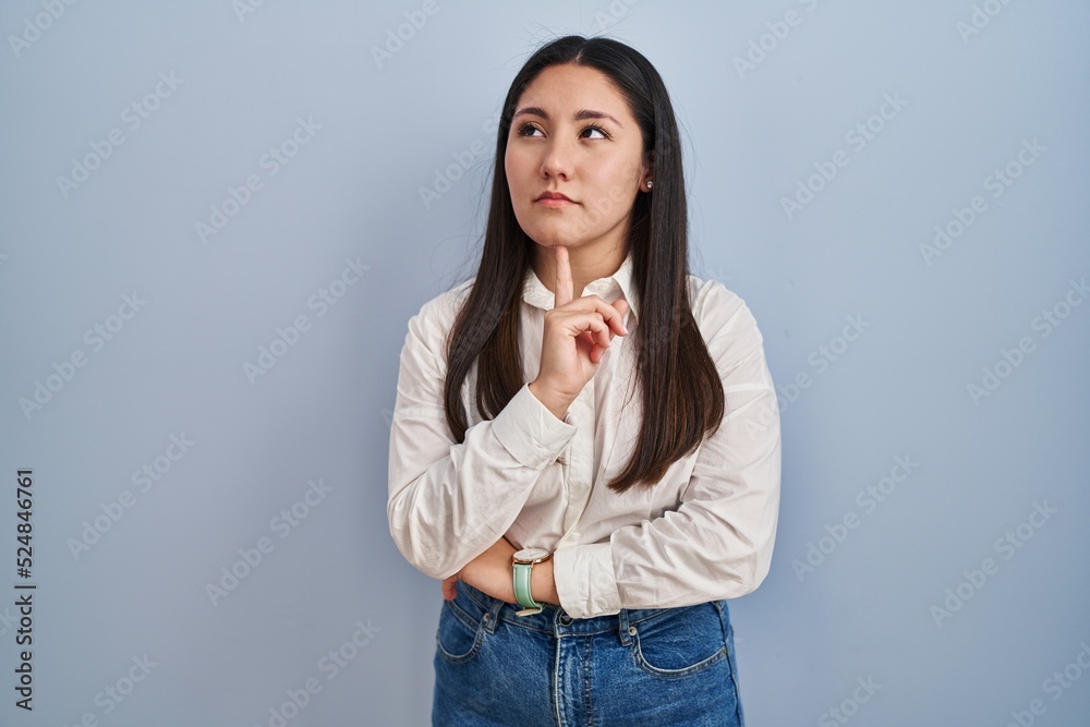 Young latin woman standing over blue background thinking concentrated about doubt with finger on chin and looking up wondering Young latin woman standing over blue background thinking concentrated about doubt with finger on chin and looking up wondering