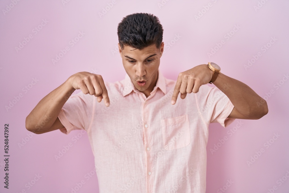 Young hispanic man standing over pink background pointing down with fingers showing advertisement, surprised face and open mouth