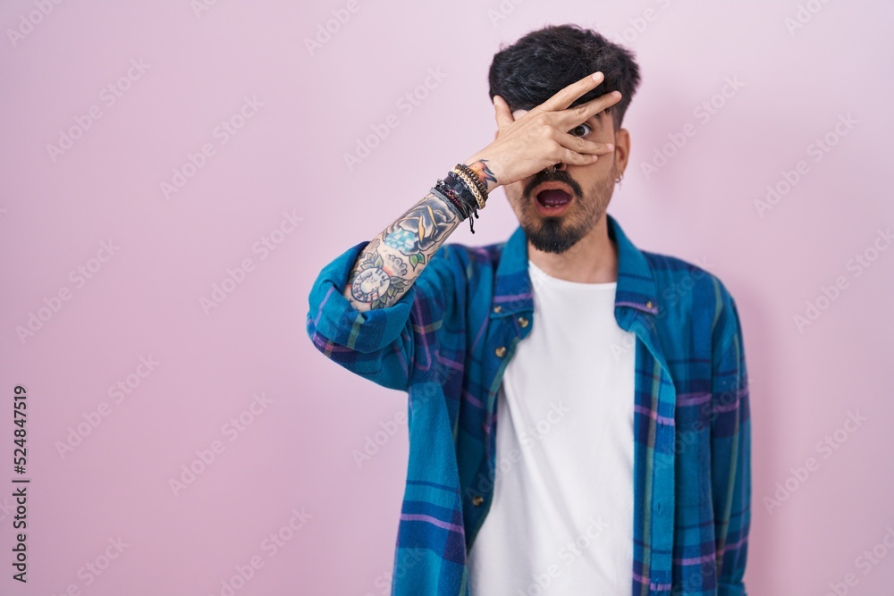 Young hispanic man with beard standing over pink background peeking in shock covering face and eyes with hand, looking through fingers with embarrassed expression.