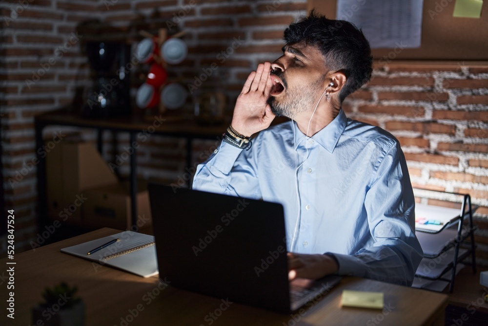 Young hispanic man with beard working at the office at night shouting and screaming loud to side with hand on mouth. communication concept. Young hispanic man with beard working at the office at night shouting and screaming loud to side with hand on mouth. communication concept.