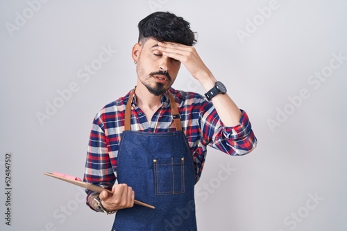 Young hispanic man with beard wearing waiter apron holding clipboard worried and stressed about a problem with hand on forehead, nervous and anxious for crisis