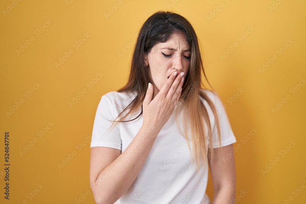 Young brunette woman standing over yellow background bored yawning tired covering mouth with hand. restless and sleepiness. Young brunette woman standing over yellow background bored yawning tired covering mouth with hand. restless and sleepiness.