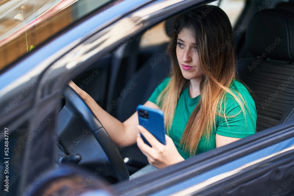 Young hispanic woman using smartphone sitting on car at street