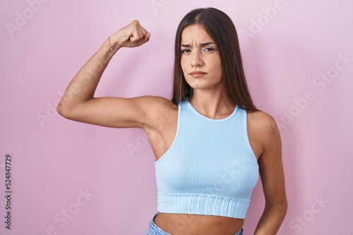 Young brunette woman standing over pink background strong person showing arm muscle  confident and proud of power
