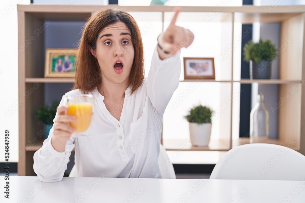 Brunette woman drinking glass of orange juice pointing with finger surprised ahead, open mouth amazed expression, something on the front Brunette woman drinking glass of orange juice pointing with finger surprised ahead, open mouth amazed expression, something on the front