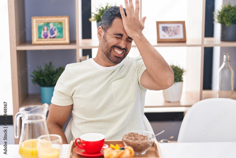 Hispanic man with beard eating breakfast surprised with hand on head for mistake, remember error. forgot, bad memory concept. Hispanic man with beard eating breakfast surprised with hand on head for mistake, remember error. forgot, bad memory concept.