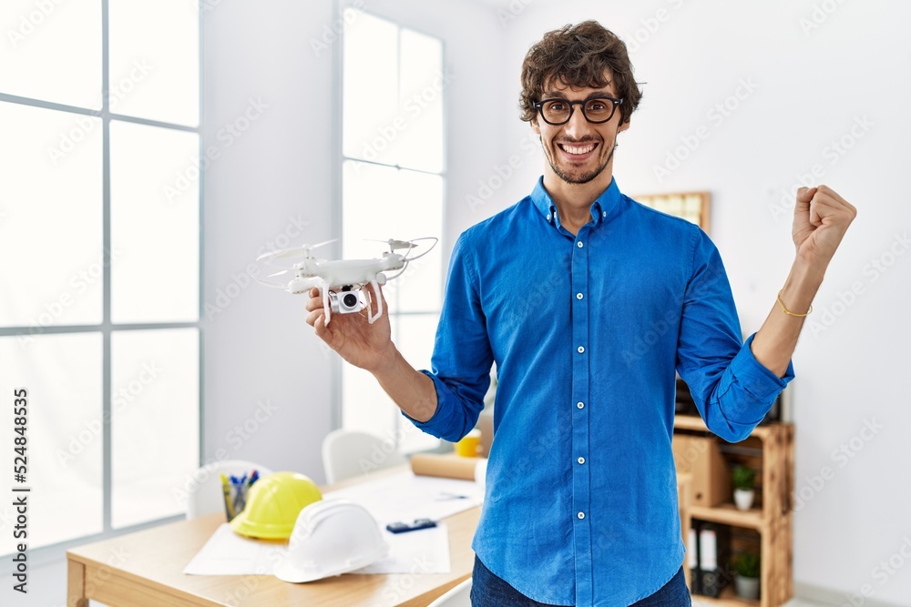 Young hispanic man using drone at architect office screaming proud, celebrating victory and success very excited with raised arm