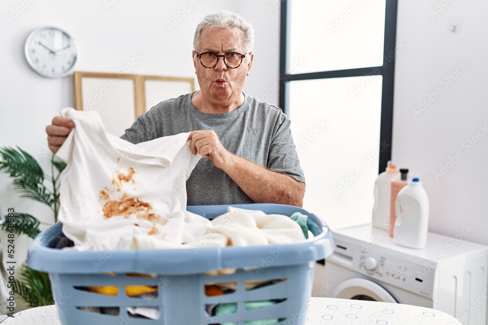 Senior caucasian man holding dirty t shirt with stain making fish face with mouth and squinting eyes, crazy and comical. Senior caucasian man holding dirty t shirt with stain making fish face with mouth and squinting eyes, crazy and comical.
