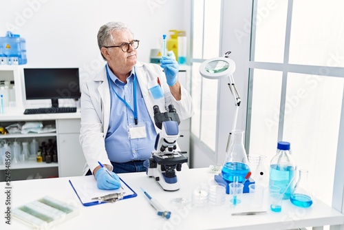 Middle age grey-haired man wearing scientist uniform holding test tube and writing on clipboard at laboratory