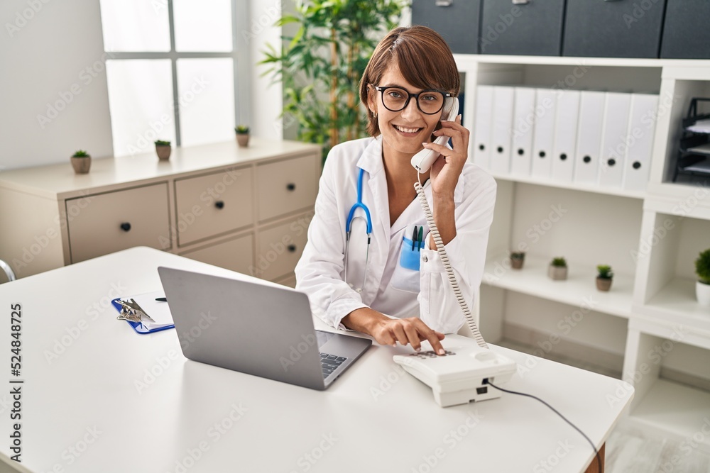 Young beautiful hispanic woman doctor using laptop talking on telephone at clinic Young beautiful hispanic woman doctor using laptop talking on telephone at clinic