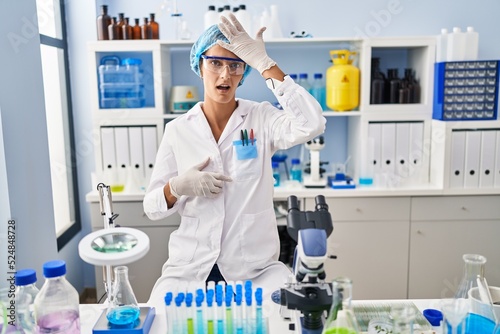 Brunette woman working at scientist laboratory surprised with hand on head for mistake, remember error. forgot, bad memory concept.
