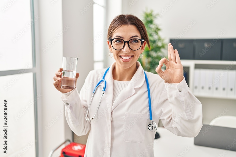 Young brunette doctor woman holding glass of water doing ok sign with fingers, smiling friendly gesturing excellent symbol Young brunette doctor woman holding glass of water doing ok sign with fingers, smiling friendly gesturing excellent symbol