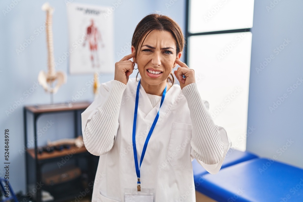 Young brunette woman working at pain recovery clinic covering ears with fingers with annoyed expression for the noise of loud music. deaf concept. Young brunette woman working at pain recovery clinic covering ears with fingers with annoyed expression for the noise of loud music. deaf concept.