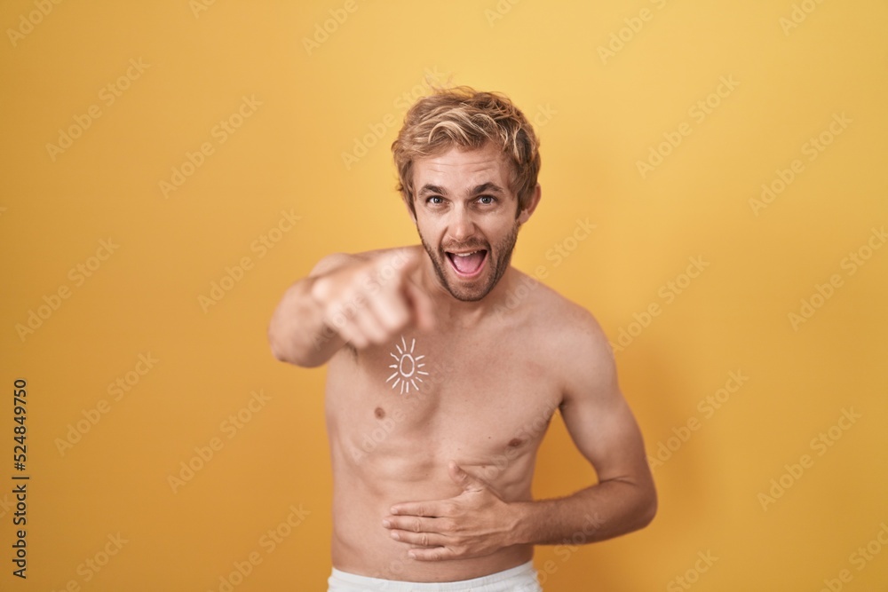 Caucasian man standing shirtless wearing sun screen laughing at you, pointing finger to the camera with hand over body, shame expression