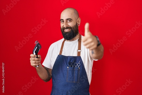 Young hispanic man with beard and tattoos wearing barber apron holding razor approving doing positive gesture with hand, thumbs up smiling and happy for success. winner gesture.