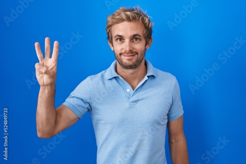 Caucasian man standing over blue background showing and pointing up with fingers number three while smiling confident and happy.