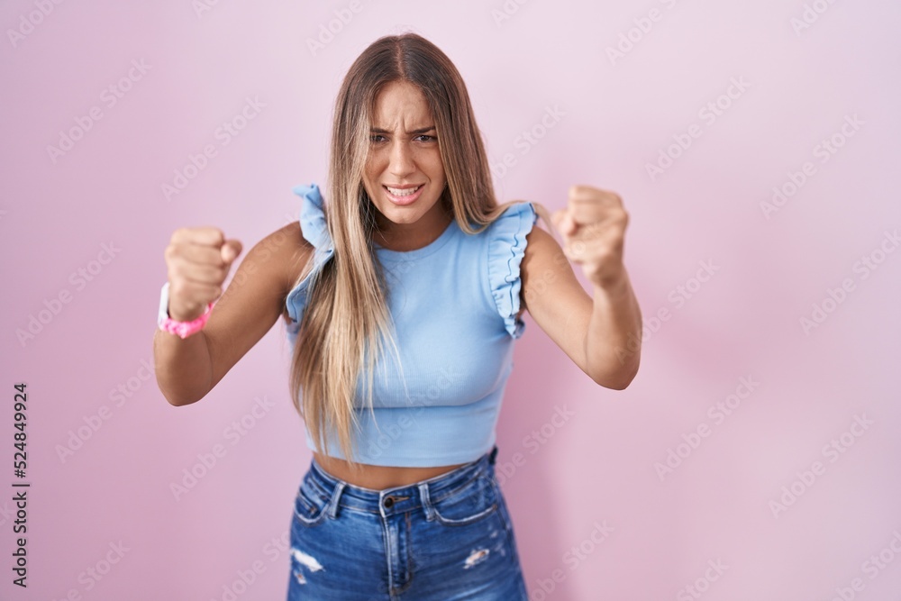 Young blonde woman standing over pink background angry and mad raising fists frustrated and furious while shouting with anger. rage and aggressive concept. Young blonde woman standing over pink background angry and mad raising fists frustrated and furious while shouting with anger. rage and aggressive concept.