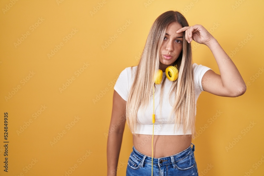 Young blonde woman standing over yellow background wearing headphones worried and stressed about a problem with hand on forehead, nervous and anxious for crisis