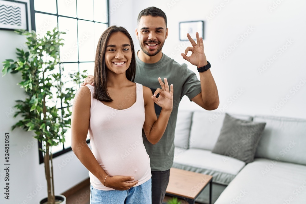 Young interracial couple expecting a baby, touching pregnant belly smiling positive doing ok sign with hand and fingers. successful expression.