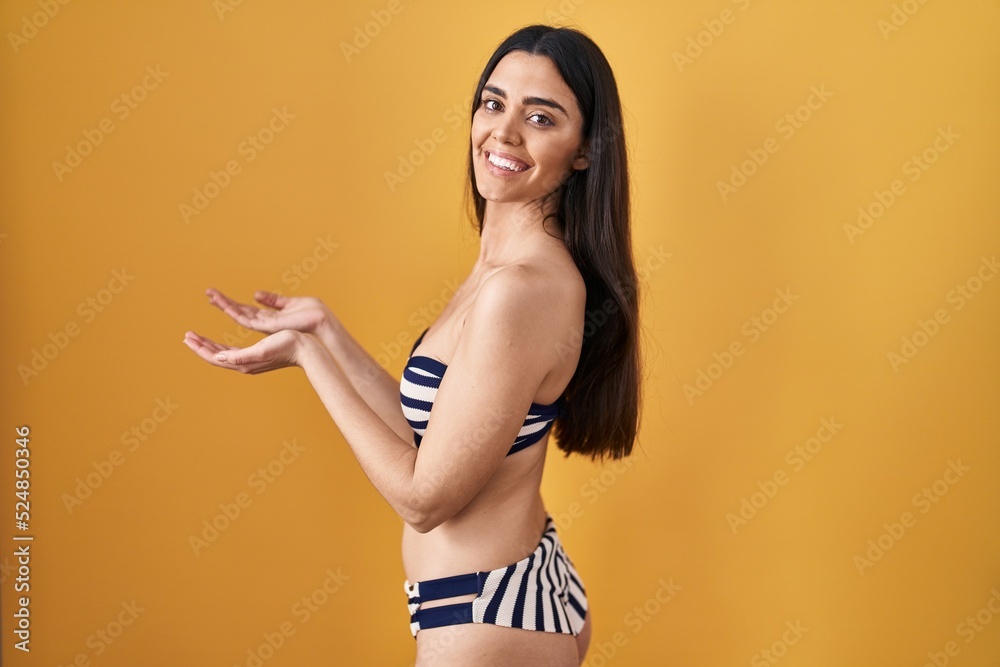 Young brunette woman wearing bikini over yellow background pointing aside with hands open palms showing copy space, presenting advertisement smiling excited happy