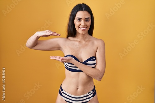 Young brunette woman wearing bikini over yellow background gesturing with hands showing big and large size sign, measure symbol. smiling looking at the camera. measuring concept.