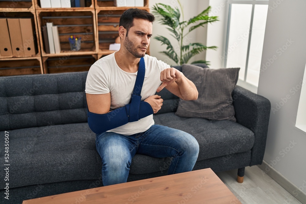 Young hispanic man with beard wearing arm on sling sitting at therapy consult with angry face, negative sign showing dislike with thumbs down, rejection concept