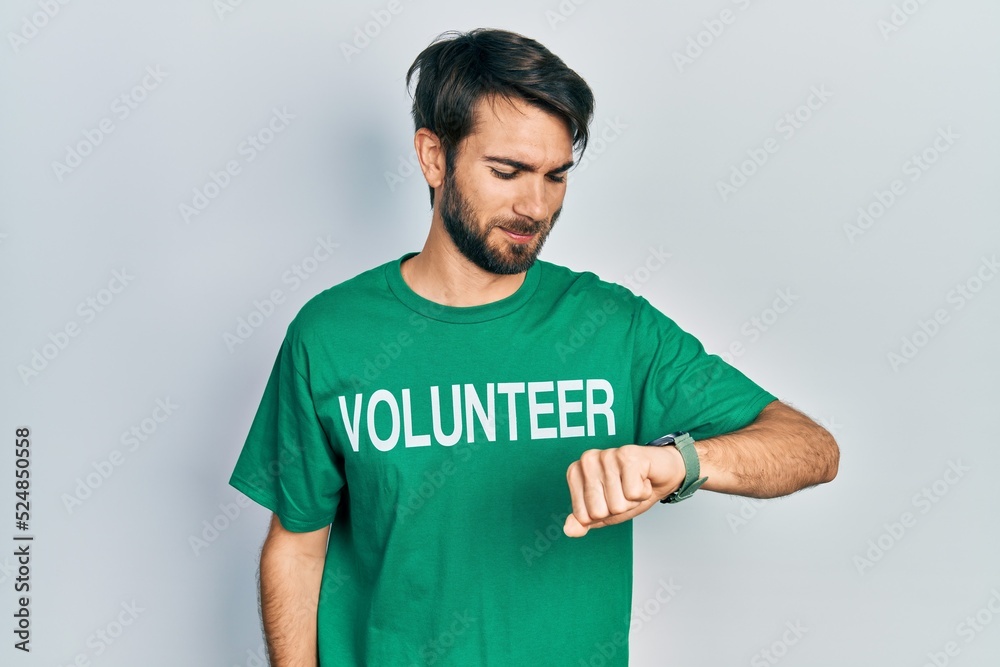 Young hispanic man wearing volunteer t shirt checking the time on wrist watch, relaxed and confident Young hispanic man wearing volunteer t shirt checking the time on wrist watch, relaxed and confident