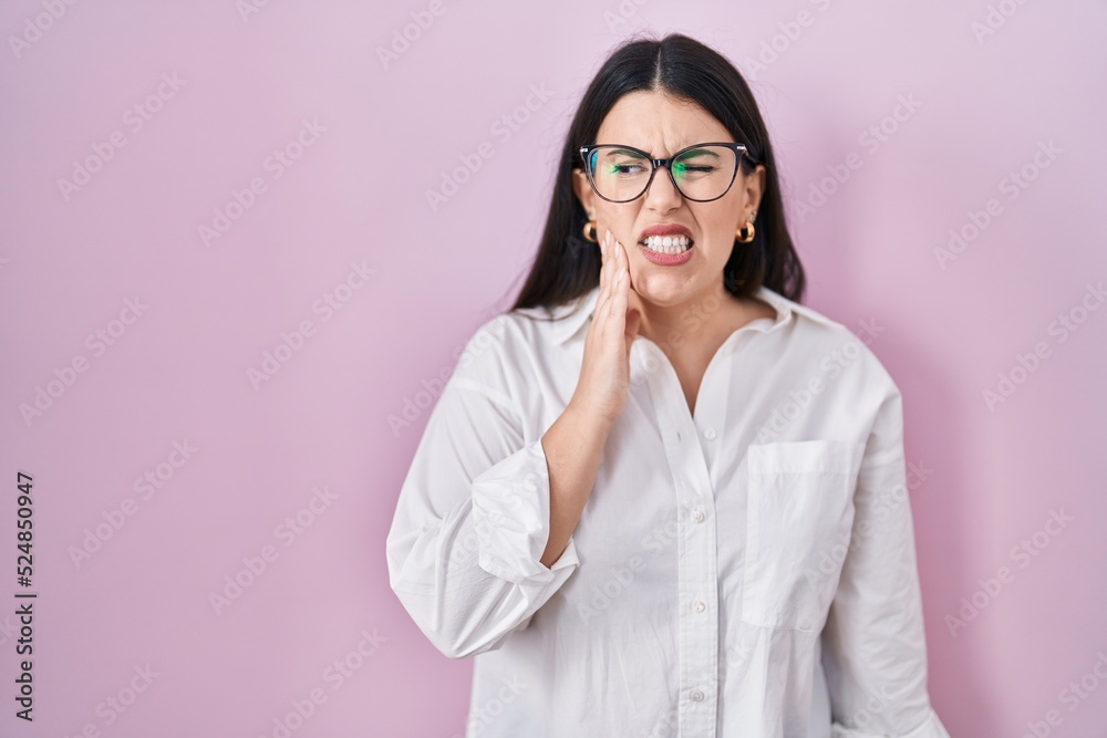 Young brunette woman standing over pink background touching mouth with hand with painful expression because of toothache or dental illness on teeth. dentist
