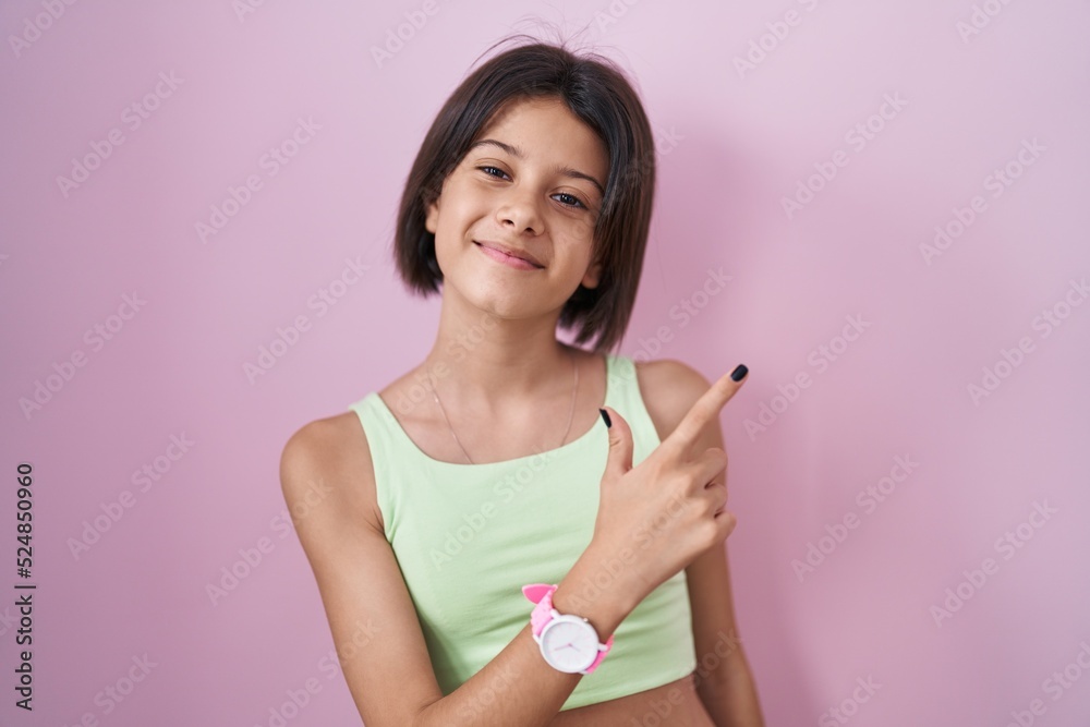Young girl standing over pink background cheerful with a smile on face pointing with hand and finger up to the side with happy and natural expression Young girl standing over pink background cheerful with a smile on face pointing with hand and finger up to the side with happy and natural expression