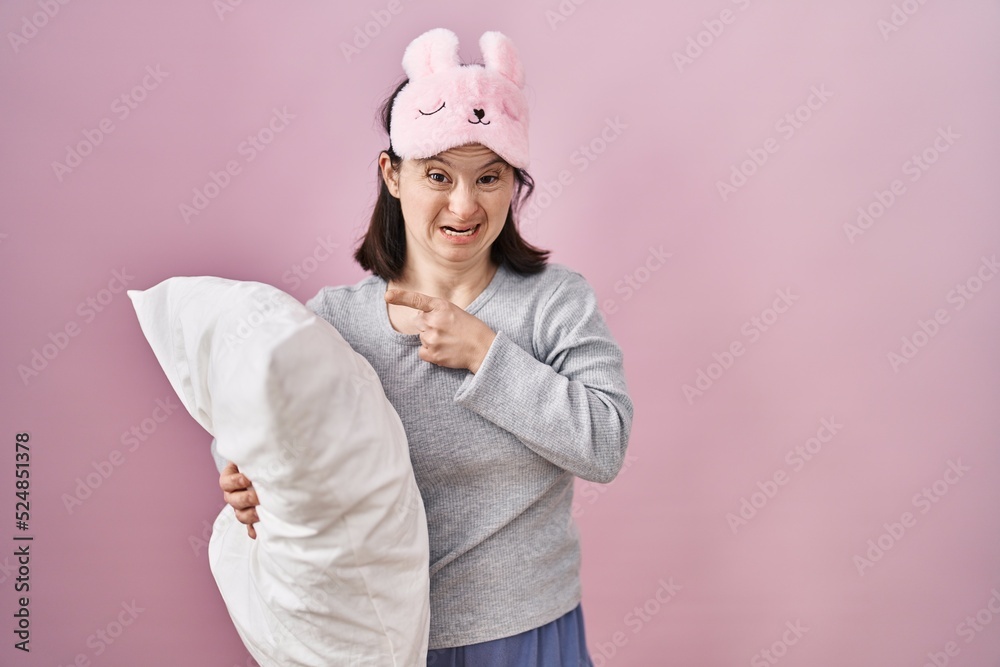 Woman with down syndrome wearing sleeping mask hugging pillow pointing aside worried and nervous with forefinger, concerned and surprised expression Woman with down syndrome wearing sleeping mask hugging pillow pointing aside worried and nervous with forefinger, concerned and surprised expression