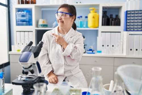 Hispanic girl with down syndrome working at scientist laboratory pointing aside worried and nervous with forefinger, concerned and surprised expression
