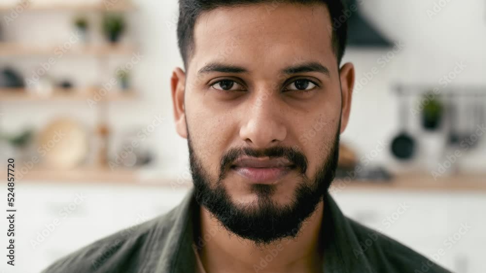 Close-up video portrait of a positive confident attractive indian or arabian young man, in casual shirt, at home in cozy living room on the background of the kitchen, looks at camera, smiles friendly
