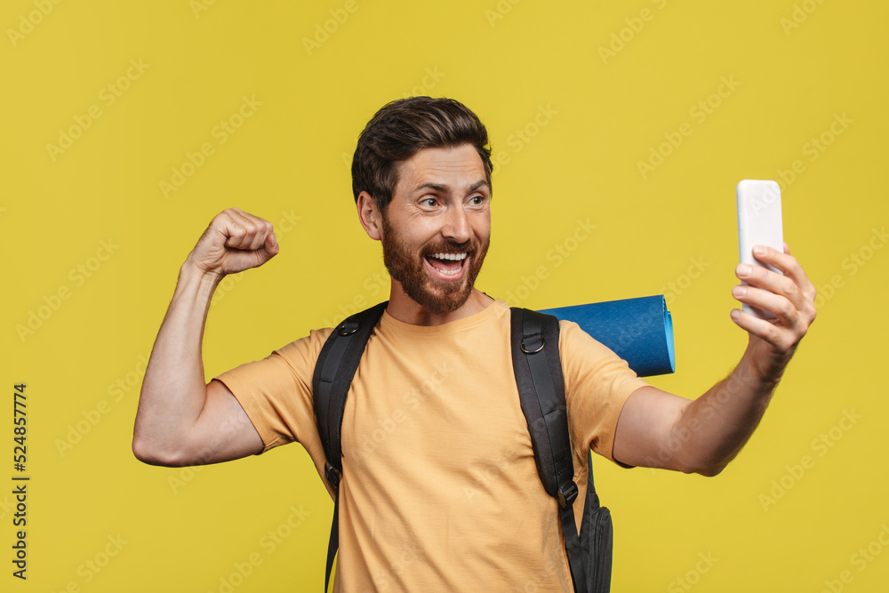 Excited man with tourist equipment taking selfie on smartphone over yellow studio background, making photo of himself Excited man with tourist equipment taking selfie on smartphone over yellow studio background, making photo of himself