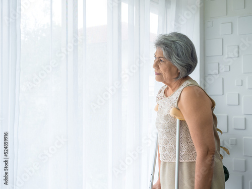 Murais de parede Asian senior woman white hair standing with walking crutches near curtain looking out glass window with copy space