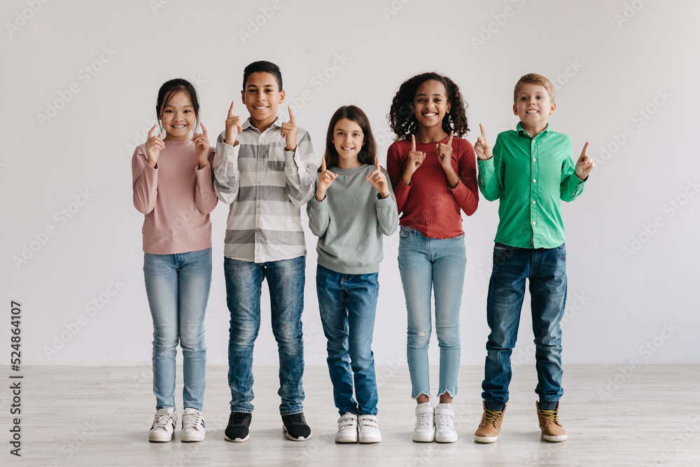 Fototapeta premium Cheerful Multicultural Preteen Children Pointing Fingers Upward Standing Together Indoors