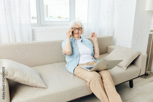 a joyful elderly lady is talking on a smartphone sitting on a sofa in a bright apartment holding a laptop on her lap smiling broadly