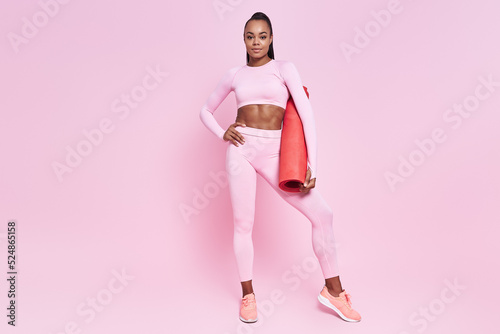 Beautiful African woman in sports clothing carrying exercise mat against pink background