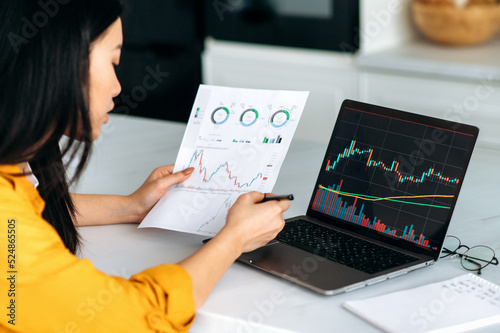 Smart successful asian female trader, stock broker, investor, analyzes growth and fall charts of cryptocurrency, online coins, predicts future performance, focused studies the market