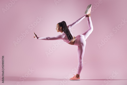 Beautiful young woman in sports clothing doing stretching exercises against pink background