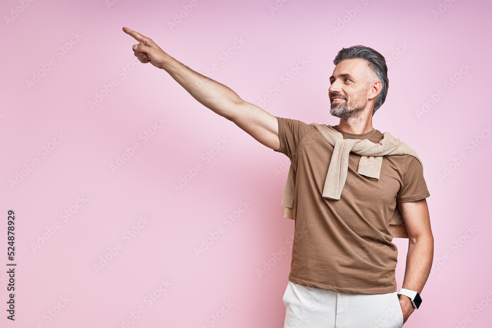 Handsome mature man pointing away and smiling while standing against pink background Handsome mature man pointing away and smiling while standing against pink background