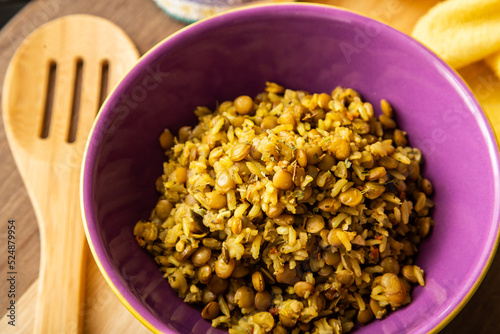 Uma vasilha de louça com arroz integral e lentilhas sobre uma superfície de madeira, com uma colher de pau e um pano amarelo ao fundo.