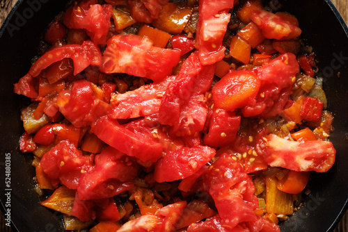 The stage of preparing a traditional shakshouka dish, adding fresh tomatoes to fried peppers with onion and garlic, close-up view